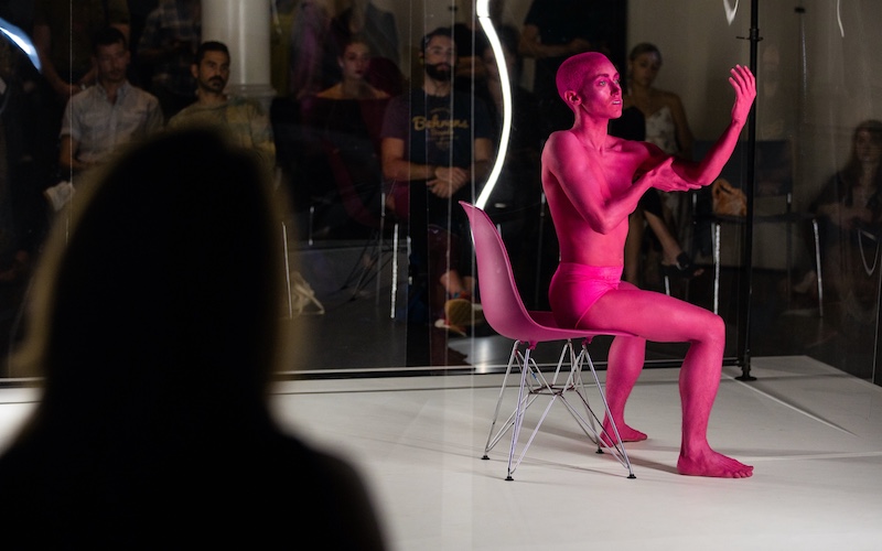 Dancer Molly Griffin sits in a hot pink office chair that resembles the shade of clay paint her body is covered in. She hold one arm out at a ninety degree angle. She holds her raised arm with her other hand by her bicep. Audience members watch behind clear plexi-glass.